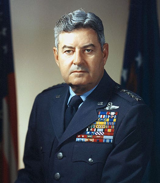 A man in military uniform with flags on the wall behind him.